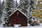 Image of a small rural church sitting amongst snow covered spruce trees, one which has a few red Christmas ornaments on it.