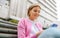 Image of smart blonde young woman wearing white t-shirt, and pink jacket, smiling and sending messages on a smartphone. Pretty