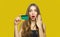 Image of surprised young lady standing over yellow background and holding debit card in hands. Looking at camera