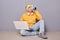 Image of thirsty man wearing beanie hat, yellow hoodie and jeans posing against gray wall, working on laptop, sitting on floor and