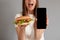 Image of unrecognizable unknown woman wearing white t-shirt holding fast food burger and showing mobile cell phone with blank