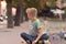 Image of young blond boy sitting cross legged looking at the street behind him with soft focus of street and tree lines