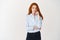 Image of young redhead female office worker biting finger and looking indecisive, having doubts or worries, standing