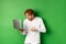 Image of young redhead man looking surprised at laptop screen, drop jaw and staring at display amazed, green background