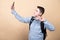 Image of youngster guy wearing casual clothing and backpack holding smartphone and taking selfie isolated over beige background