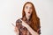 Impressed young woman with red long hair and pale skin, staring amazed and pointing fingers left at advertisement