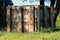 Improvised homemade compost box made of wooden pallets at the garden edge surrounded with uncut grass and tall tree in background