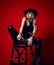 Impudent blonde woman in black leather pants, top and stetson hat sits on stepladder holding her feet in hand
