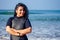 Indian female surfer dressed in black wet suit is standing near water with crossed hands