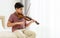 Indian handsome teenage boy, playing, practicing violin musical instrument with happiness in bedroom at cozy home in leisure time