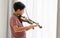 Indian handsome teenage boy, playing, practicing violin musical instrument with happiness in bedroom at cozy home in leisure time