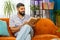 Indian man reading interesting book turning pages smiling enjoying literature taking a rest on sofa