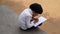 Indian schoolgirl is sitting on the stone in the park and writing in a book. A little girl is doing her homework in nature