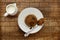 An individual spotted dick sponge cake, on a white plate with a spoon, and a small jug of cream, on a checked wooden surface with