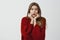 Indoor portrait of worried timit european female coworker in red loose sweater, holding hands on chin and dropping jaw