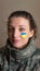 Indoor portrait of young girl with blue and yellow ukrainian flag on her cheek wearing military uniform, mandatory