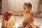 Indoor shot of adorable toddler girl with pigtails wearing yellow striped dress, playing with doll while sitting on floor in sunny