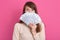 Indoor shot of astonished woman standing against pink studio wall, covering her face with fan of money, posing with big eyes,