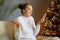 Indoor shot of beautiful woman wearing white sweater sitting on sofa near Christmas tree and having video call or broadcasting