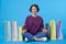 Indoor shot of cheerful young pretty dark haired curly woman keeping her legs crossed while posing over blue background with her