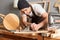 Indoor shot of concentrated young carpenter wearing apron and cap working in workshop, using plane, making wooden chair, enjoying