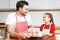 Indoor shot of handsome man wears striped apron and his small pretty female kid hold hand made cakes, rejoices