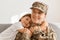 Indoor shot of happy positive military woman wearing camouflage uniform and cap, returning home from war or army, sitting on sofa