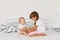 Indoor shot of happy sibling girls sitting on bed on light bedroom and playing together, elder sister spending time with infant