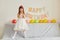 Indoor shot of happy smiling female child posing at home on her birthday, standing on sofa in white elegant dress and clapping