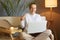 Indoor shot of joyful optimistic beautiful woman wearing white jumper sitting on cough and working on laptop or watching film,