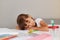 Indoor shot of little schoolgirl sleeping while sitting at the table, being tired while doing doing her homework, child with dark