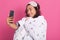 Indoor shot of lovely young woman sleep mask and wrappedwhite blanket, taking selfie during bedtime against pink background,