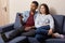 Indoor shot of multiethnic couple watch television at home on comfortable sofa. Black man holds remote control, switches on TV,