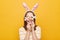 Indoor shot of playful smiling woman dressed rabbit ears covering her eyes with Easter cake pops isolated over yellow background,