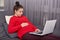 Indoor shot of pregnant female dressed red sweater and maroon leggings, keeps hand on belly, watches films online, uses wireless