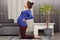 Indoor shot of pregnant lady with hair bun, has big belly, keeps hands on back, dressed in casual clothing, stands on knees,