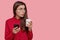 Indoor shot of puzzled surprised dark haired beautiful woman holds take out coffee, modern cell phone, dressed in red