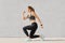 Indoor shot of self determined woman with pony tail, stands on one knee, warms up before cardio training, wears tanktop, leggings,