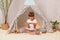 Indoor shot of shocked little girl with pigtails wearing white t shirt posing in wigwam at home and using phone, looking at