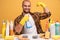 Indoor shot of smiling bearded man points at muscle, carries sponge and cleanser, brings house to order,  over yellow