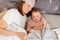 Indoor shot of smiling beautiful young mother with her little daughter reading book while laying in bed, attractive female