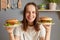 Indoor shot of smiling cheerful Caucasian woman holding two delicious fresh burgers, posing in kitchen, breaking diet, feels