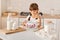 Indoor shot of smiling little cute female child with dark hair wearing white t shirt cooking in light kitchen, sitting at table