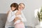 Indoor shot of smiling woman hugging her daughter, family expressing love and happiness, being glad to spend time together at