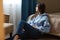 Indoor shot of thoughtful dark haired woman dressed in blue sweatshirt, jeans, sits on floor near bed, looks thoughtfully at