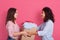 Indoor shot of two attractive women posing isolated over rosy background, smiling lady handing big carton box with clothes, young