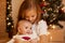 Indoor shot of two sisters posing near Christmas tree at home, elder girl hugging infant baby with love, merry Christmas and happy