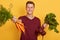 Indoor shot of young beautiful handsome gardener wearing maroon t shirt, showwing bunch of carrot to camera, attractive male
