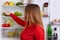 Indoor shot of young female with long straight dark hair, puts vegetables on shelf of refrigerator, eats only healthy food. Woman