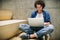 Indoor shot of young man with curly hair using laptop for chatting online with friends, browsing Internet, sitting on the stairs